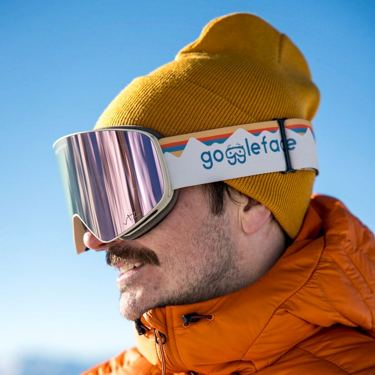 A person wearing a mustard yellow beanie and reflective snowboard goggles stands against a clear blue sky. The person is also sporting an orange jacket, and the goggle strap reads "Goggleface." Ready for any winter adventure, they showcase their Straight Line | Alpine Retro goggles with style.
